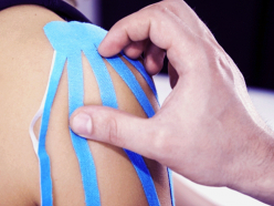 Schulter einer Frau von der Seite mit blauem Taping, Therapeut drückt es fest